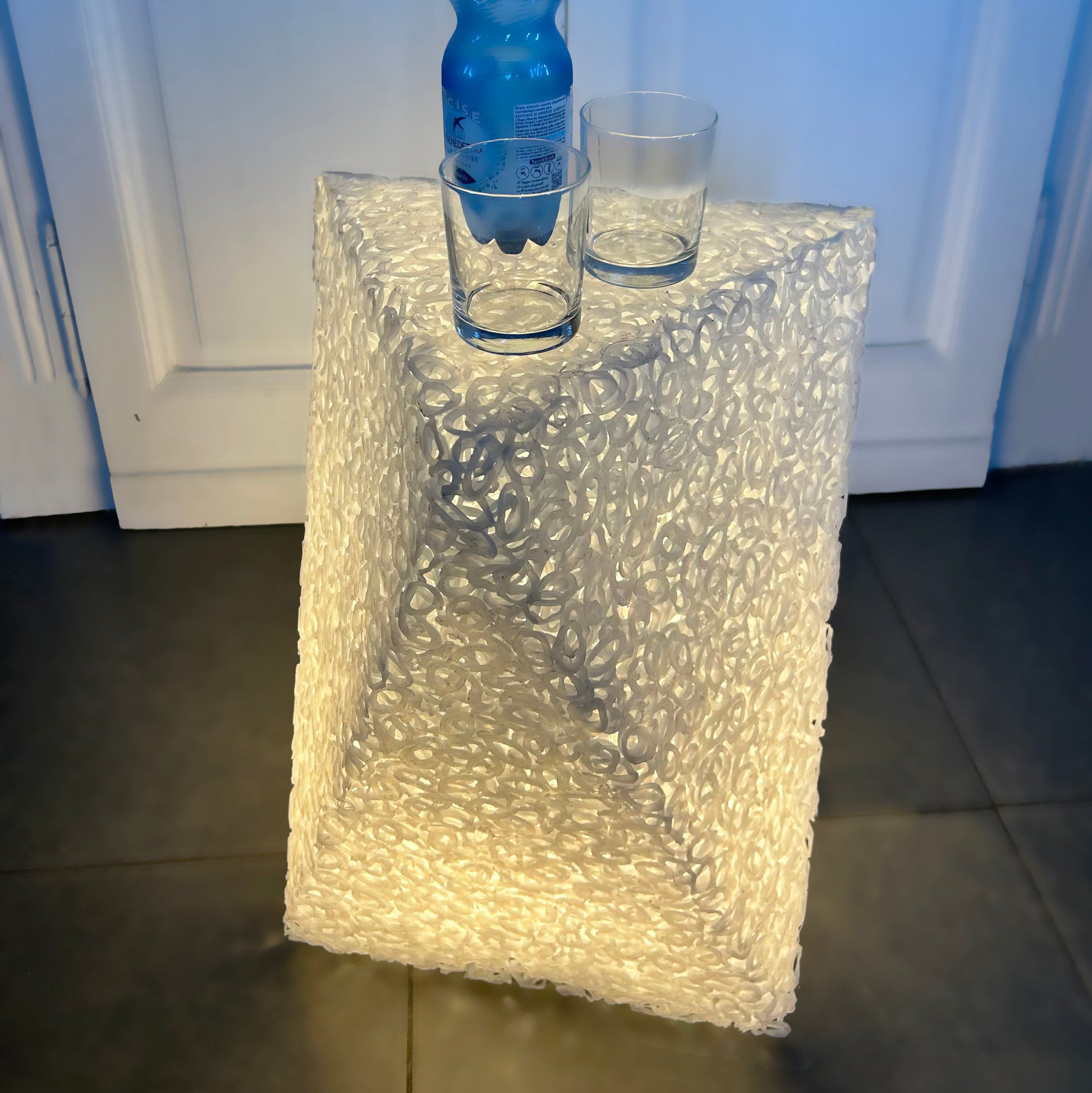 A clear, textured bubble-glass or acrylic table lamp with a geometric, faceted design, illuminated from within to cast a warm glow. A blue water bottle and two clear drinking glasses are placed on top of the lamp, which is set on a dark tiled floor in front of white paneled doors.