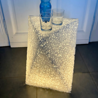 A clear, textured bubble-glass or acrylic table lamp with a geometric, faceted design, illuminated from within to cast a warm glow. A blue water bottle and two clear drinking glasses are placed on top of the lamp, which is set on a dark tiled floor in front of white paneled doors.