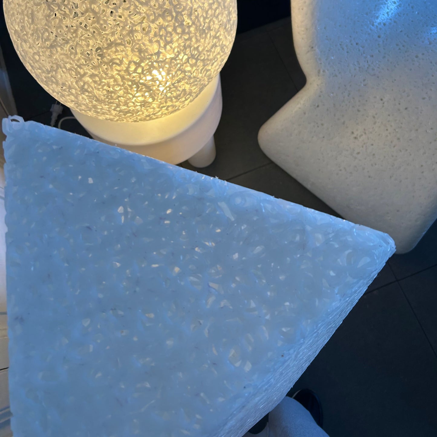 Three modern sculptural side tables with textured stone-like finishes in white, beige, and light blue-gray, shown in a dark room setting.