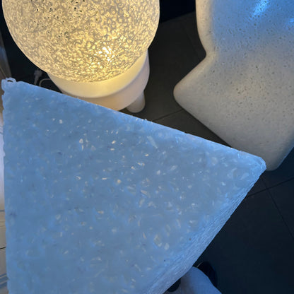 Three modern sculptural side tables with textured stone-like finishes in white, beige, and light blue-gray, shown in a dark room setting.