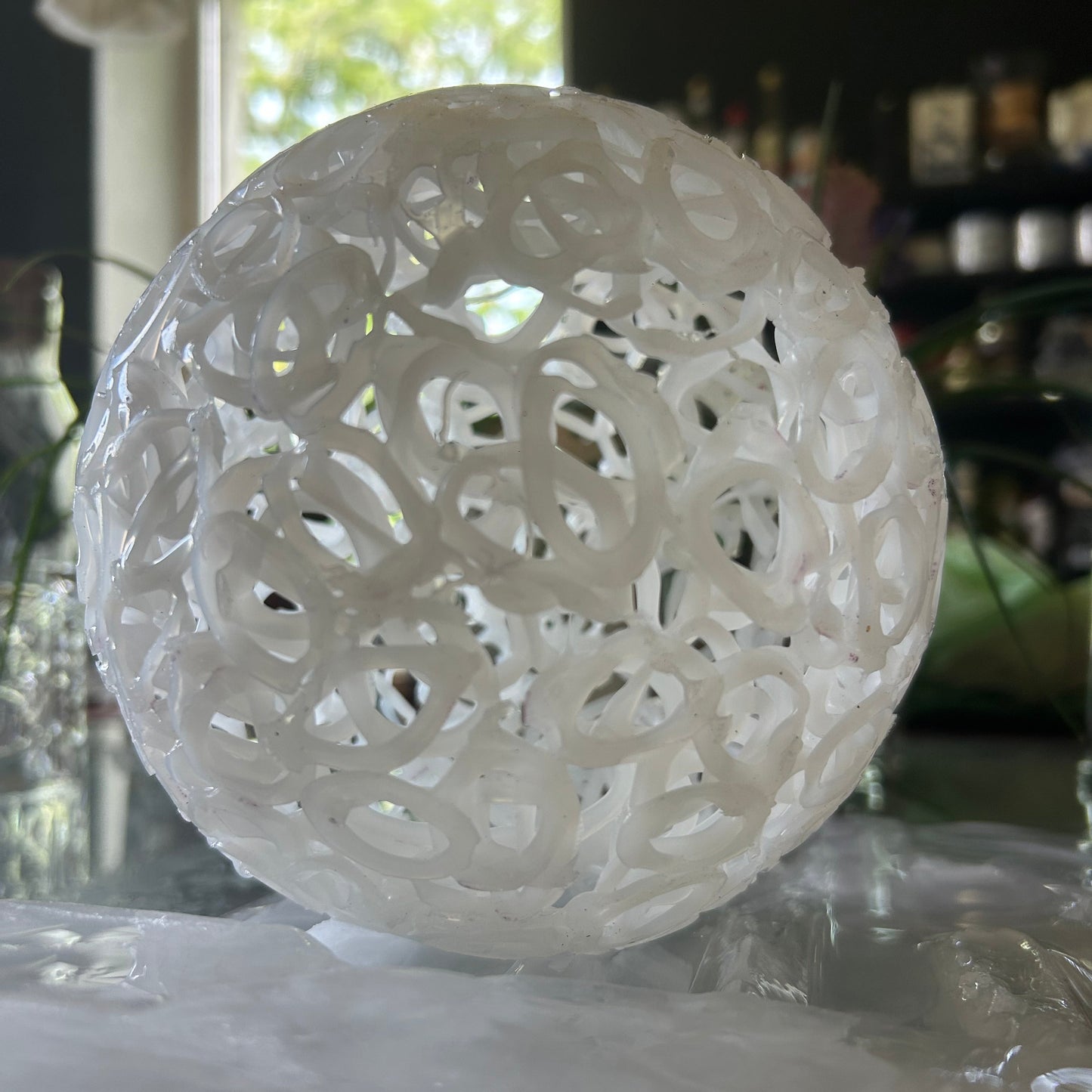 A white, spherical, open-lattice decorative sculpture or lampshade made of interlocking circular rings, shown indoors against a reflective glass surface.