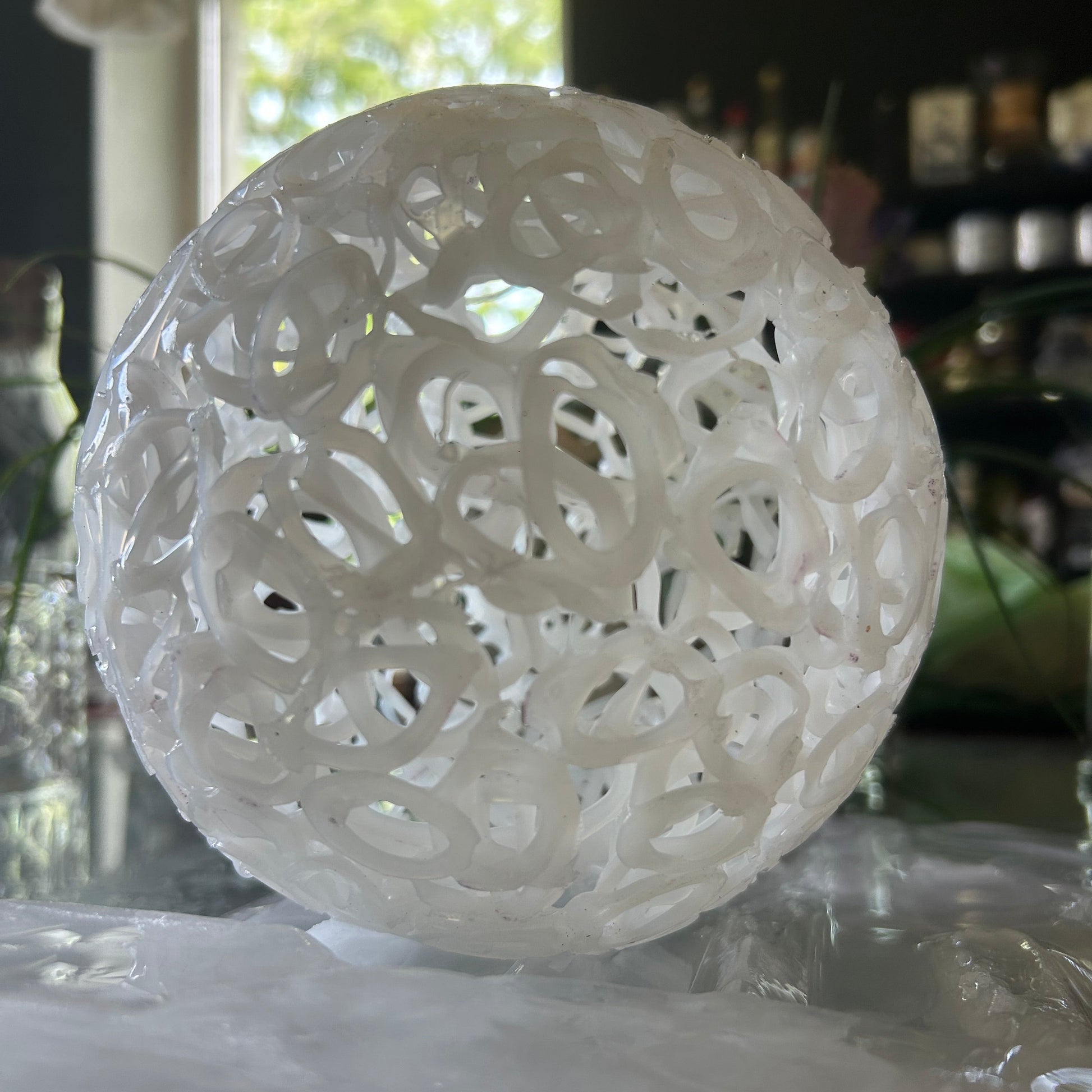 A white, spherical, open-lattice decorative sculpture or lampshade made of interlocking circular rings, shown indoors against a reflective glass surface.