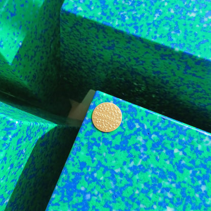 A close-up of a small round gold coin placed on a textured green and blue speckled surface made of geometric blocks.