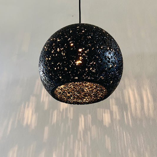 A spherical black pendant light fixture made with recycled plastics, ecopixel-open pixel, suspended by a thin black cord against a pale green wall with light reflections below.