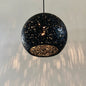 A spherical black pendant light fixture made with recycled plastics, ecopixel-open pixel, suspended by a thin black cord against a pale green wall with light reflections below.