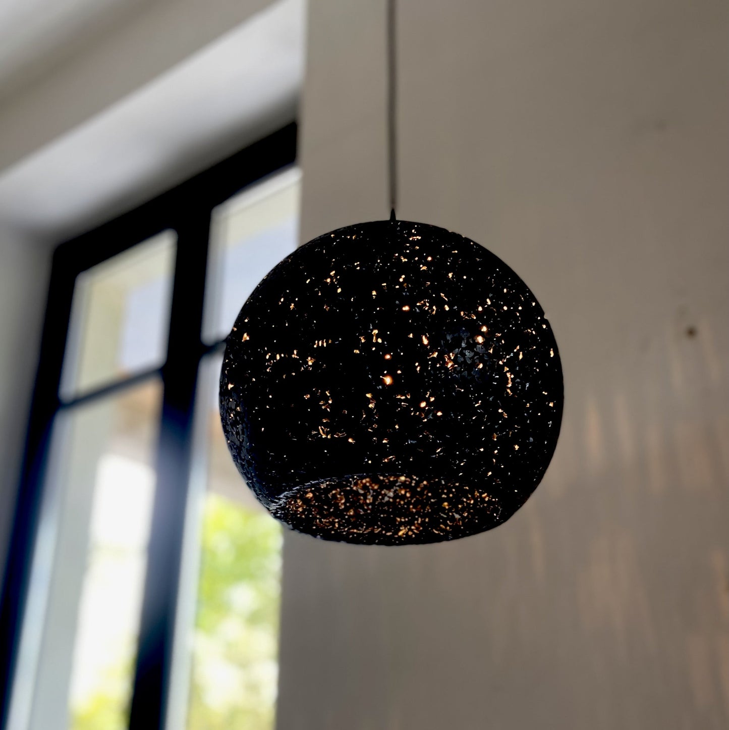 A spherical black pendant light with a textured, glittery speckled pattern hangs from a thin cord in an indoor and light-colored wall in the background.