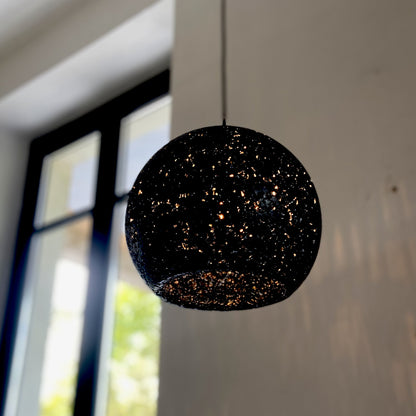 A spherical black pendant light with a textured, glittery speckled pattern hangs from a thin cord in an indoor and light-colored wall in the background.
