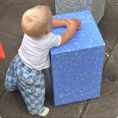 A toddler stands on a stone pavement, leaning on a blue yonder speckled cube table from ecopixel recycled plastics. Courtesy @2323Coffe Sheffield 
