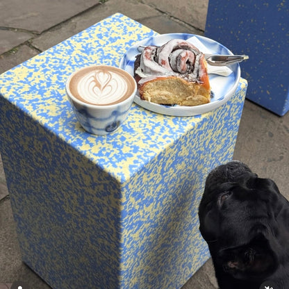 A blue and yellow speckled cube side table in an outdoor patio setting, holding a cup of latte with art and a plate of food, with a black dog's head visible in the lower right corner.