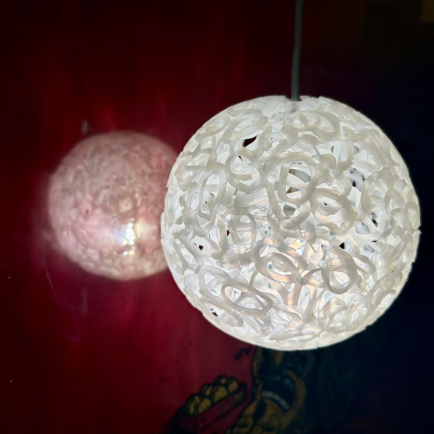 A white spherical pendant light with an intricate openwork filigree pattern, glowing with a warm light against a dark red background. A second, similar but unlit light is visible in the background.