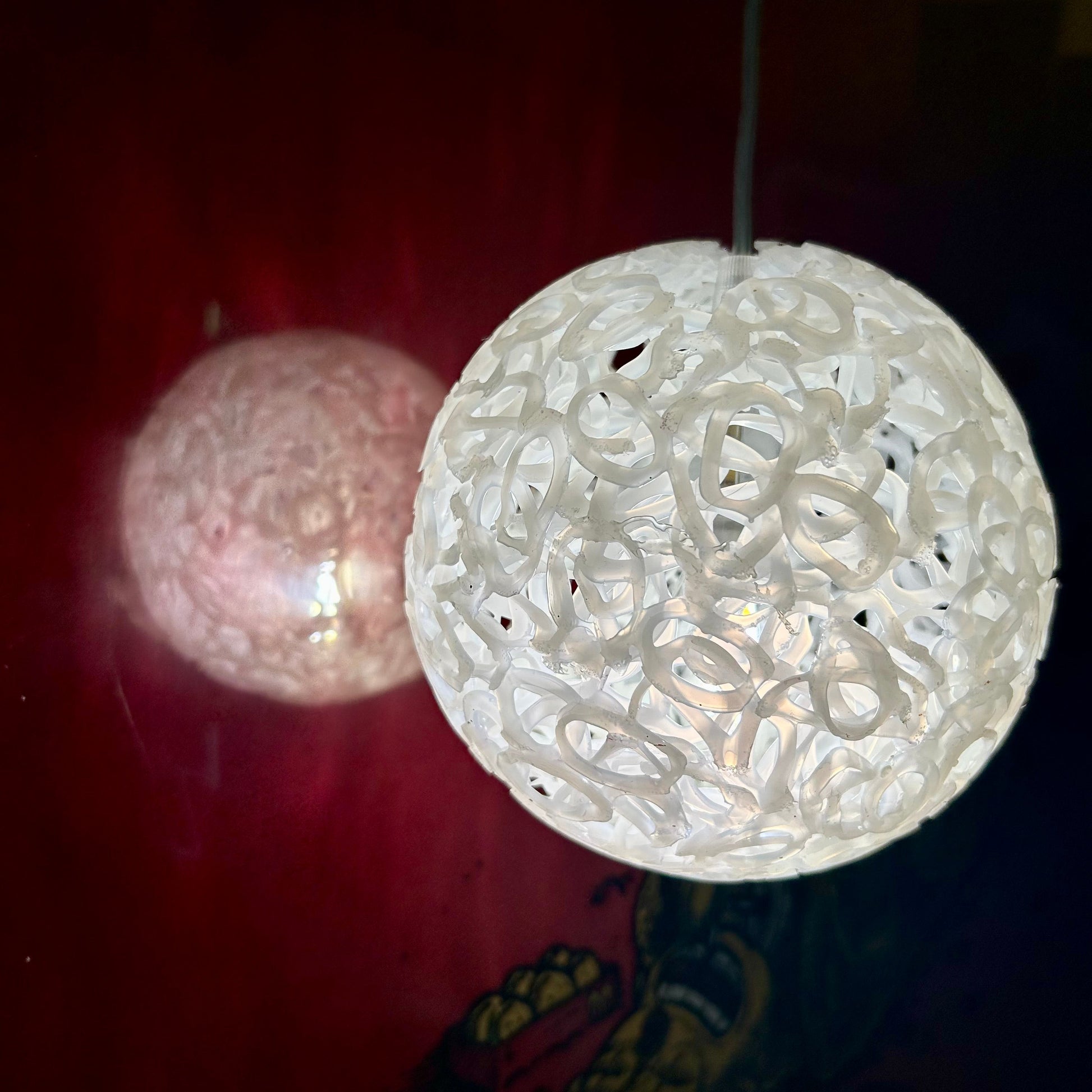 A white spherical pendant light with an intricate openwork filigree pattern, glowing with a warm light against a dark red background. A second, similar but unlit light is visible in the background.